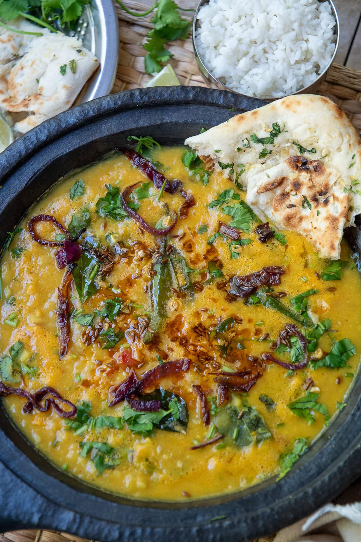 Dal tadka served in a black bowl along with a piece of naan. It is flanked by a small bowl of white rice and wedges of lime along with strands of cilantro.