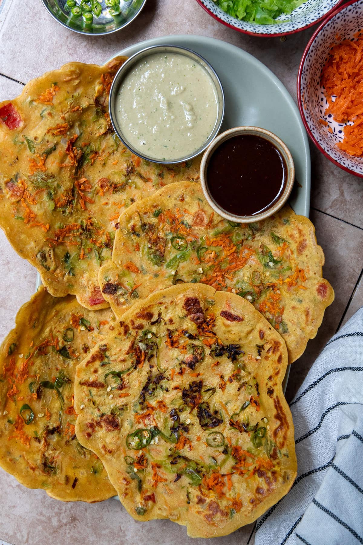 Besan Chillas are served alongside chutneys and flanked by bowls of grated carrots, bell pepper and some chopped green chilis.